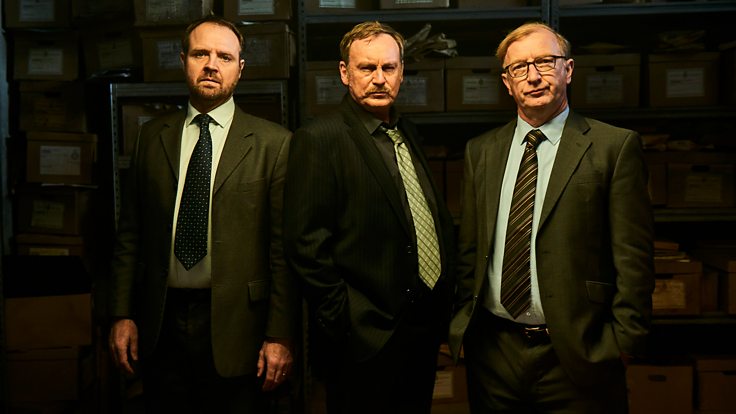A photo showing actors Gareth John Bale as DC Geraint Bale, Philip Glenister as DCI Paul Bethell and Steffan Rhodri as Phil Bach Rees stood in front of boxes of case files