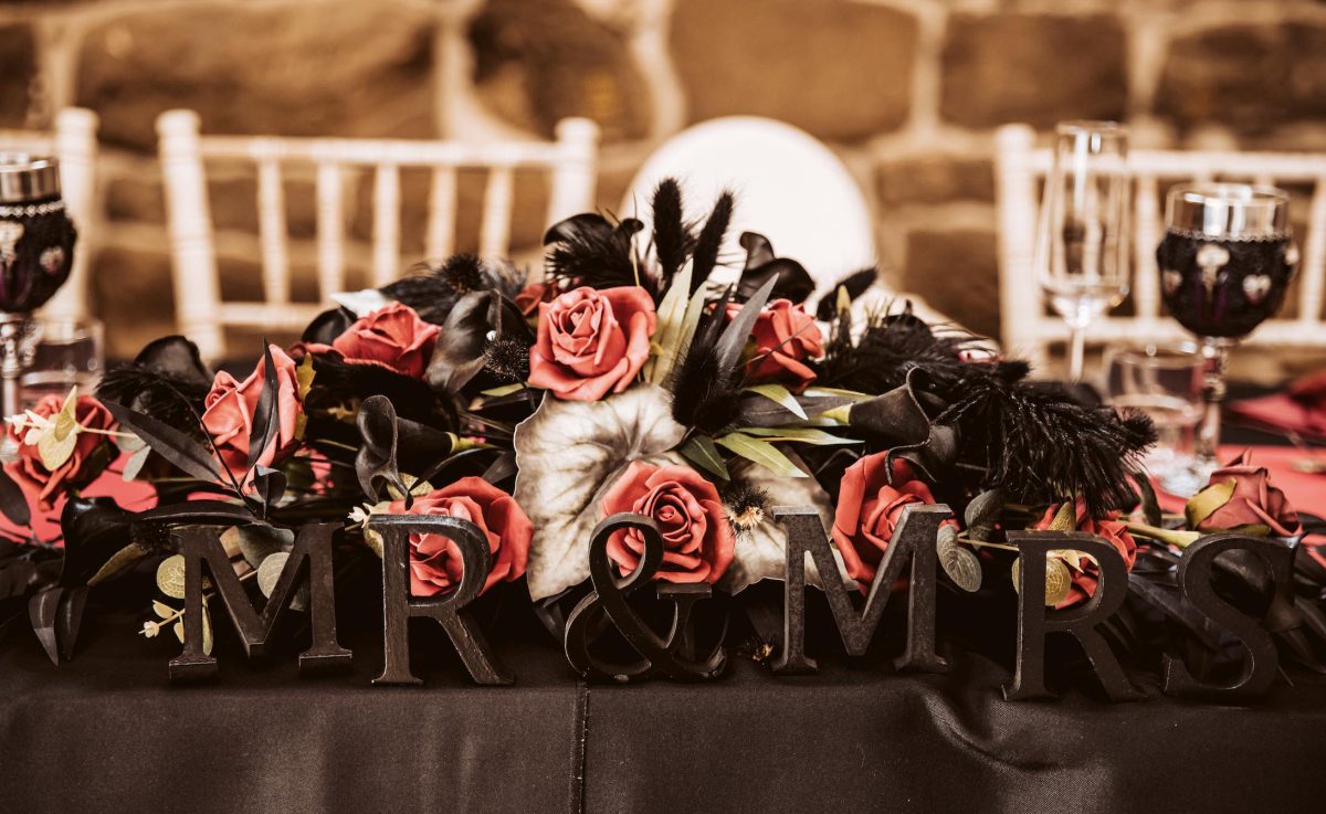 A photo of a floral table display, there are red roses and black feathers and some black wooden blocks that read Mr & Mrs in front of the display